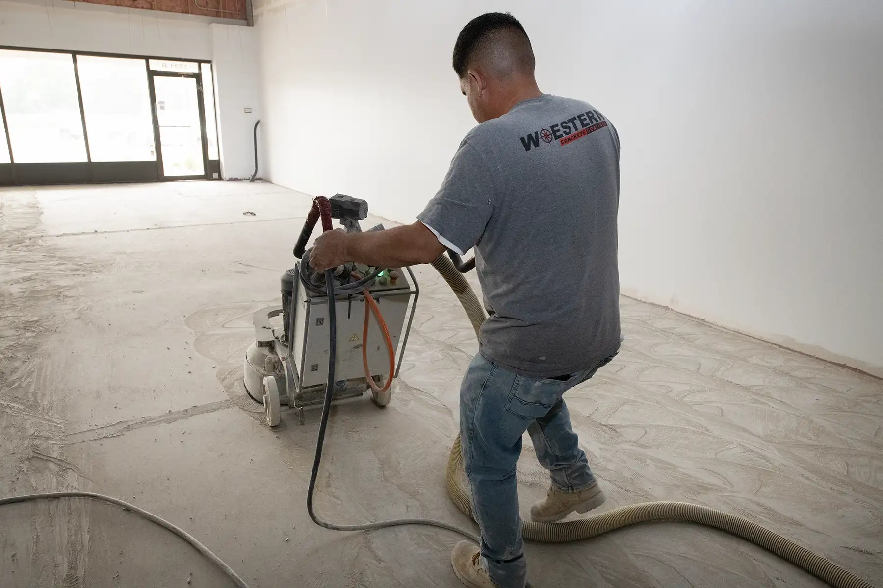 A Western Concrete Coatings crew member in a company shirt grinding a concrete floor in a bright storefront space