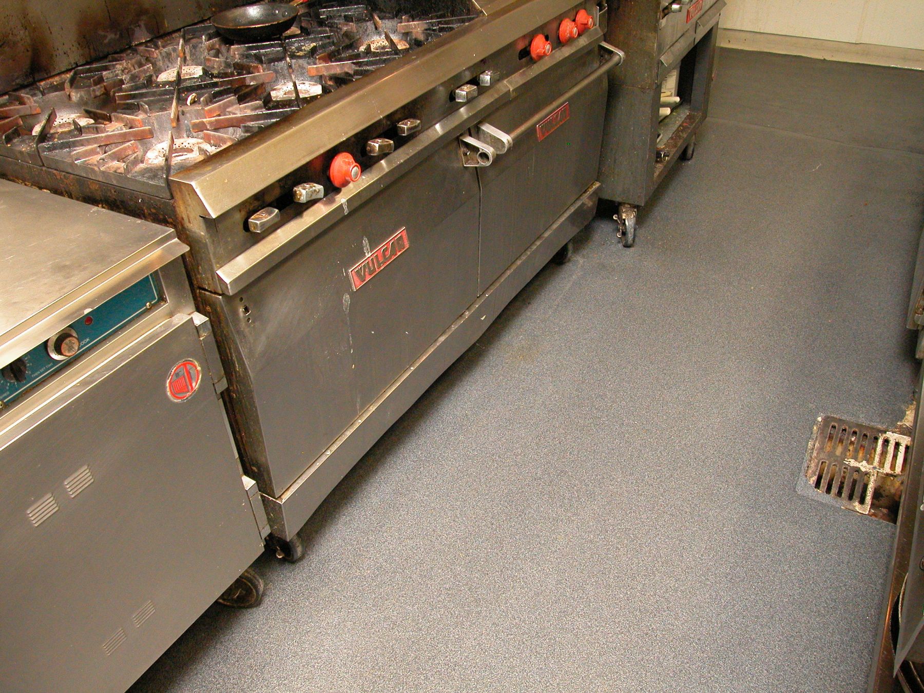 Gray speckled epoxy floor between a worn commercial gas range and kitchen equipment near a floor drain