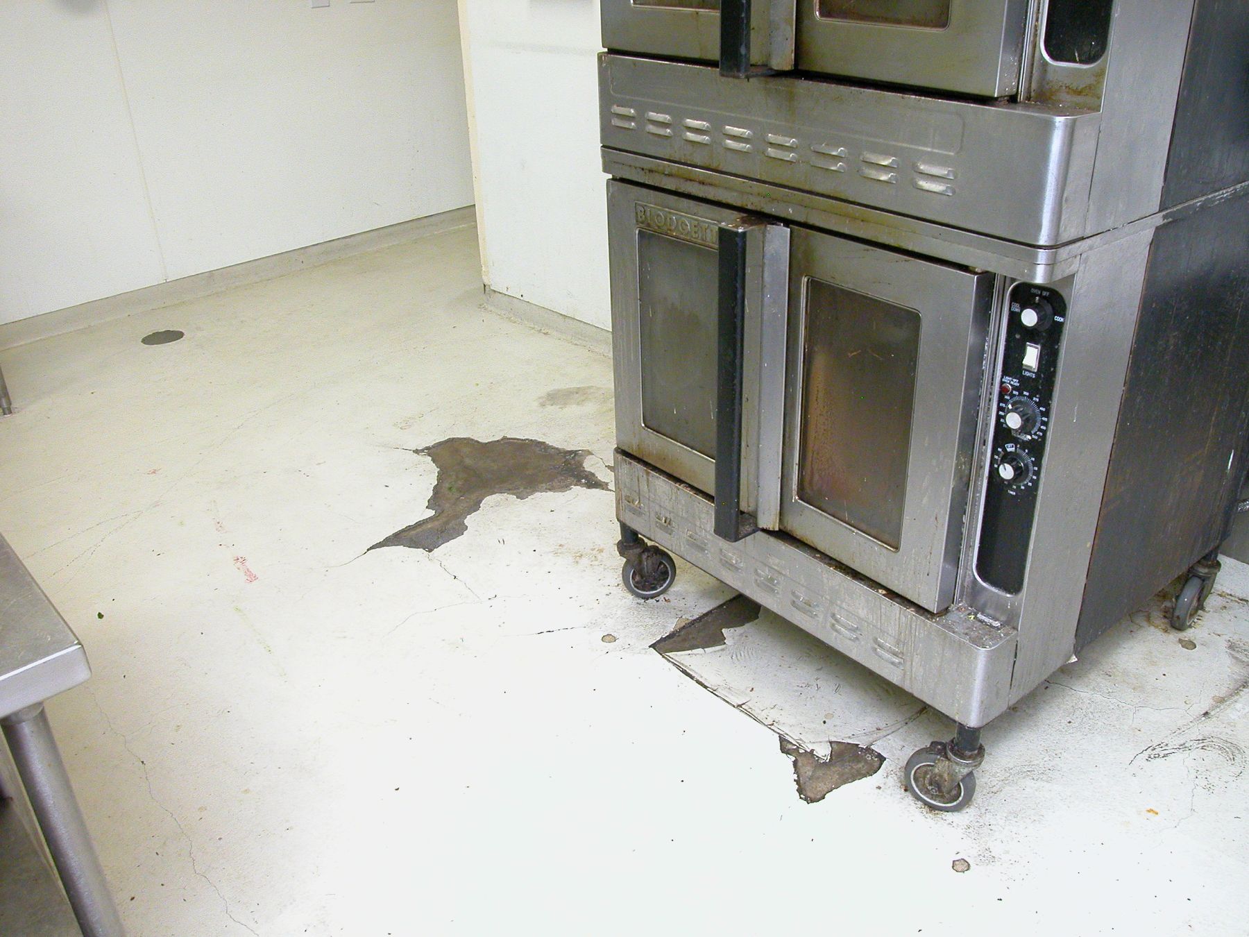 Before photo of cracked, peeling white kitchen floor coating with exposed gray patches beside a commercial convection oven