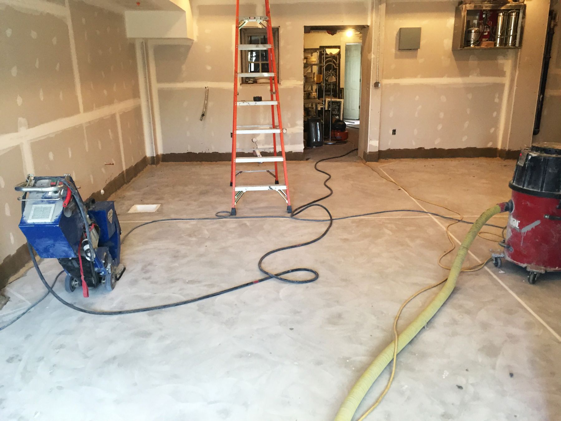 Bare ground concrete floor in a gutted commercial kitchen with grinder, vacuum and ladder
