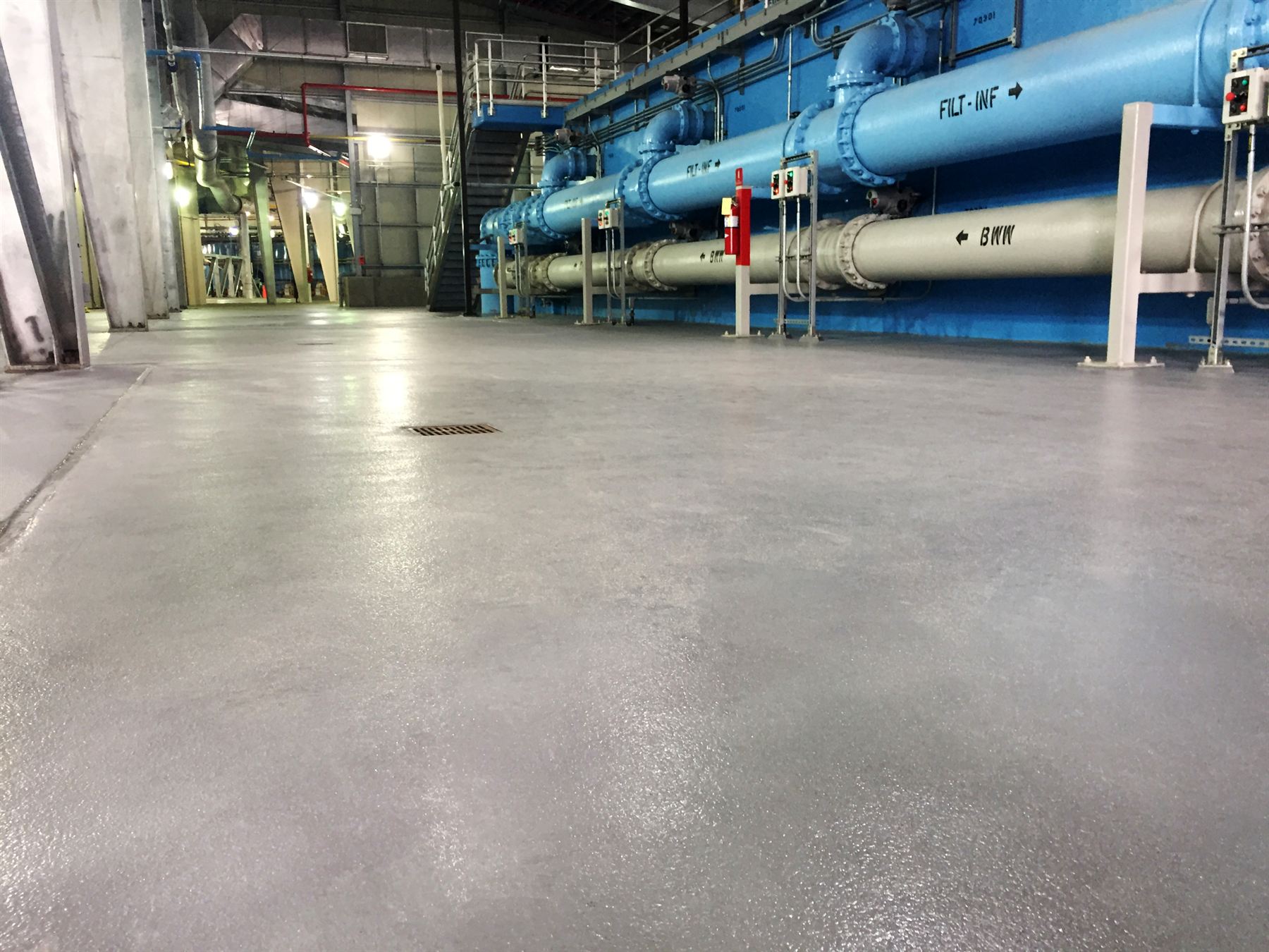 Gray textured concrete coating with floor drain in a water treatment plant beside blue pipes