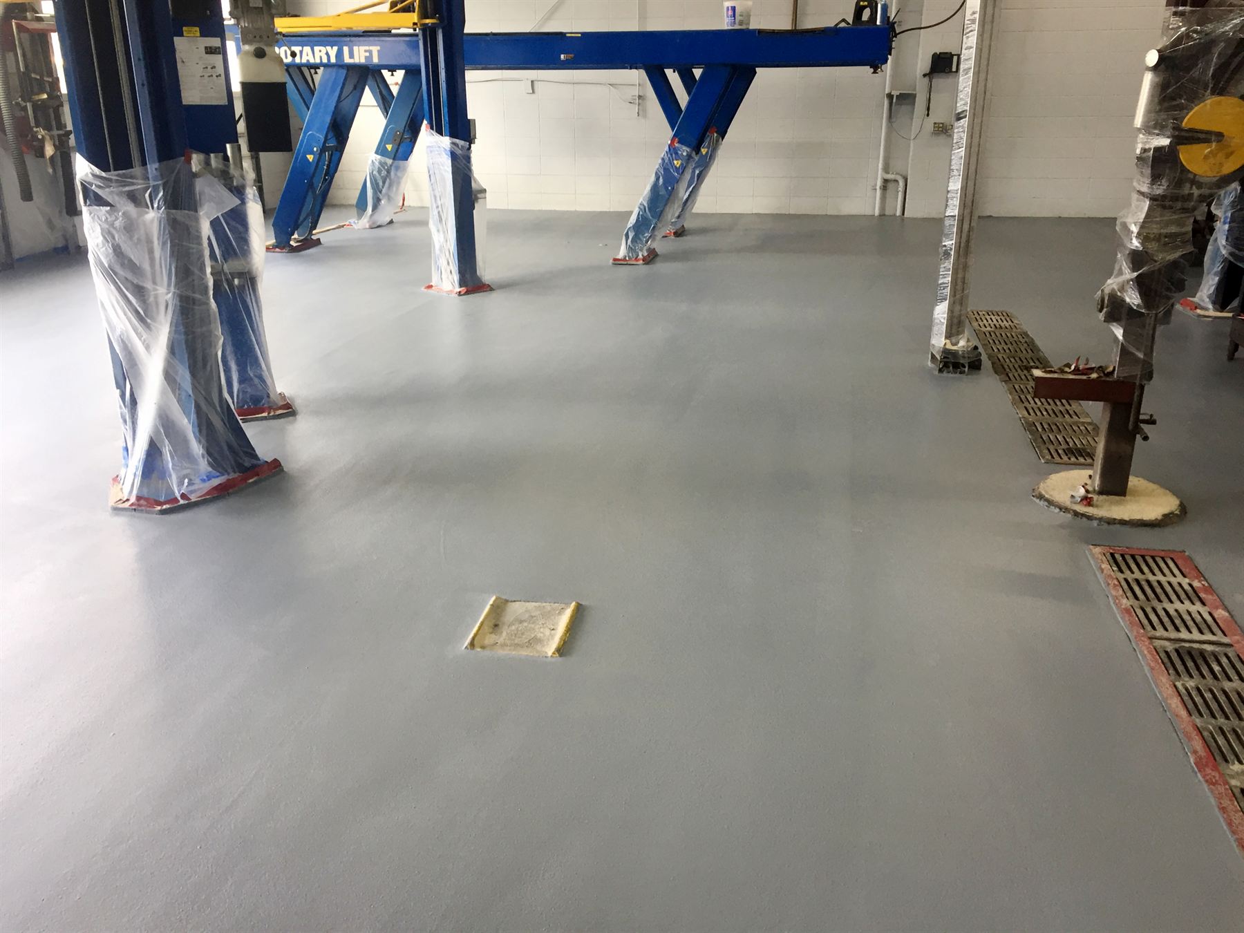 Smooth gray epoxy-coated auto shop floor around plastic-wrapped blue vehicle lifts and drain grates