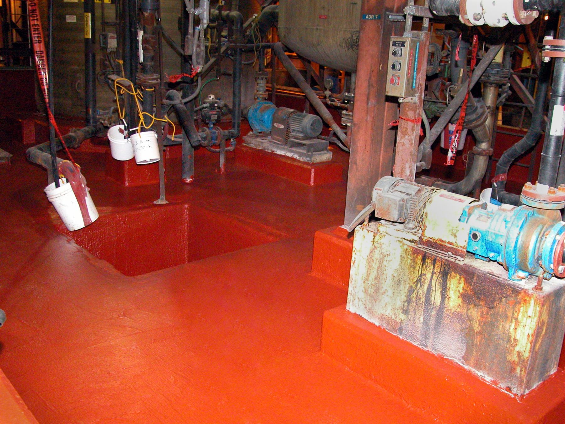 Red epoxy-coated containment floor and sump pit beside rusted pump equipment in a chemical processing plant