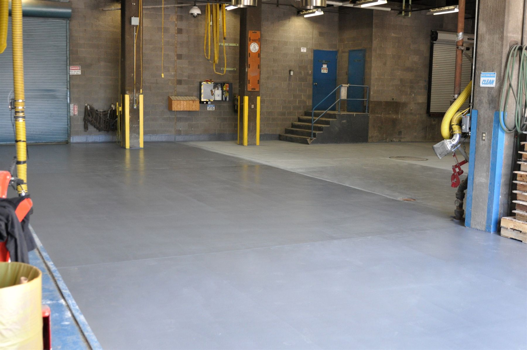 Smooth gray industrial epoxy floor coating across a loading bay with block walls, yellow bollards and blue doors