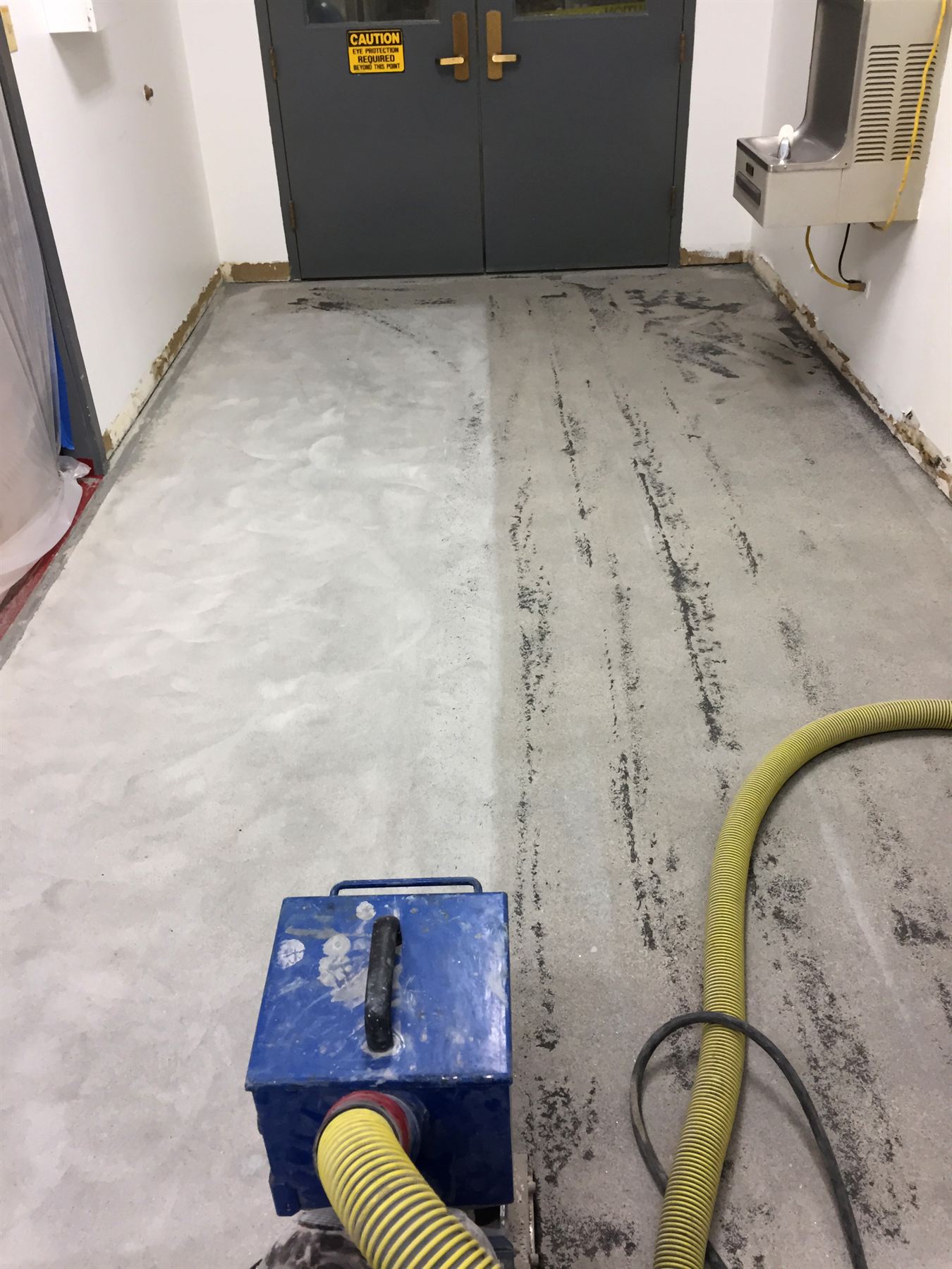 Concrete hallway floor half ground bare with a blue grinder and yellow dust hose before coating