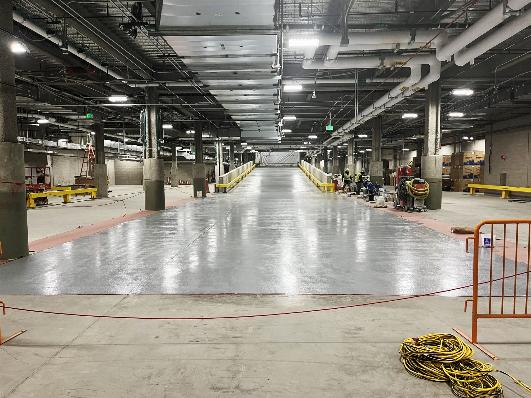 Glossy gray waterproofing coating applied to a long concrete floor lane in a large commercial building with overhead piping